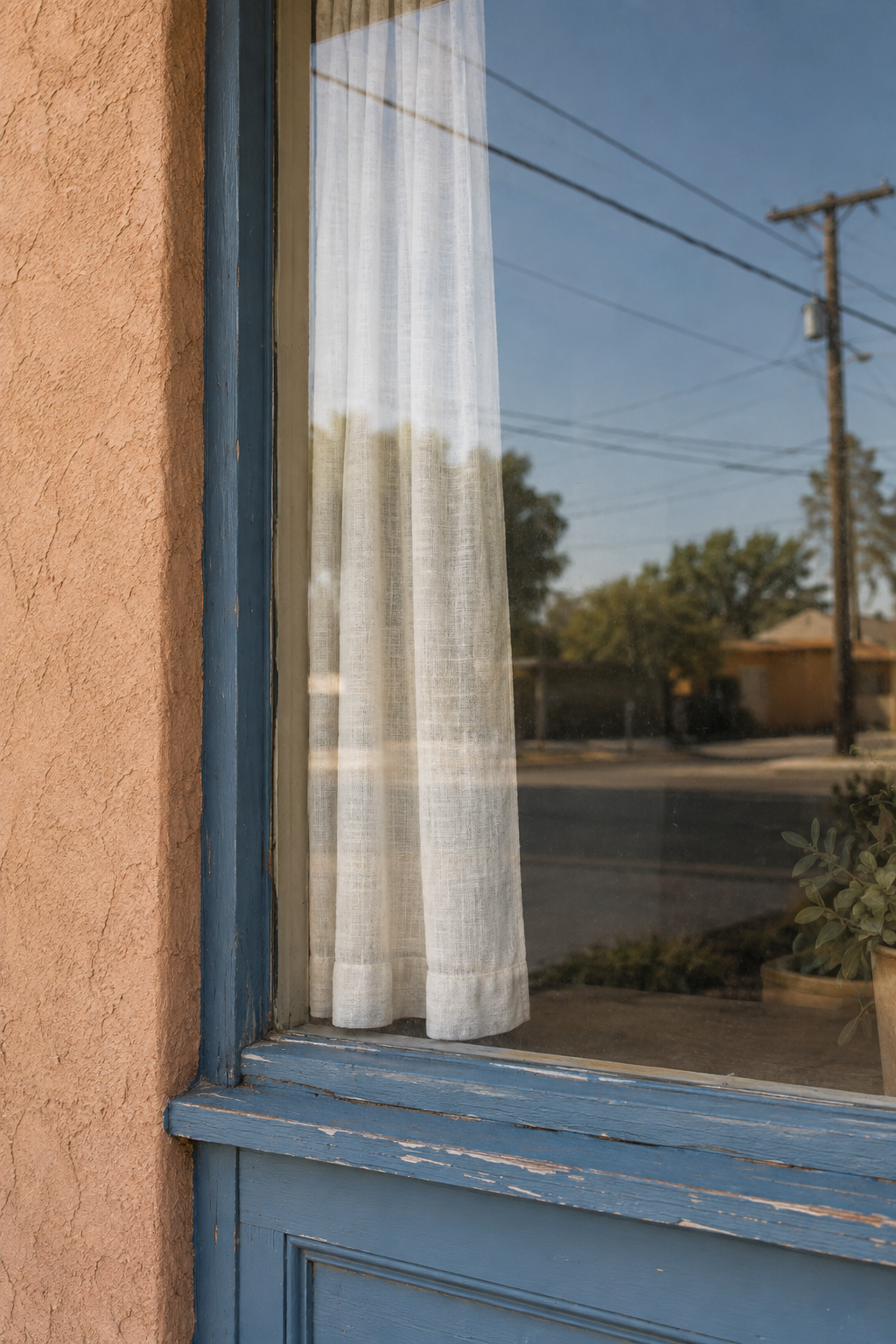 A blue-framed store window reflecting a pale sky and neighborhood wires over a cream curtain.