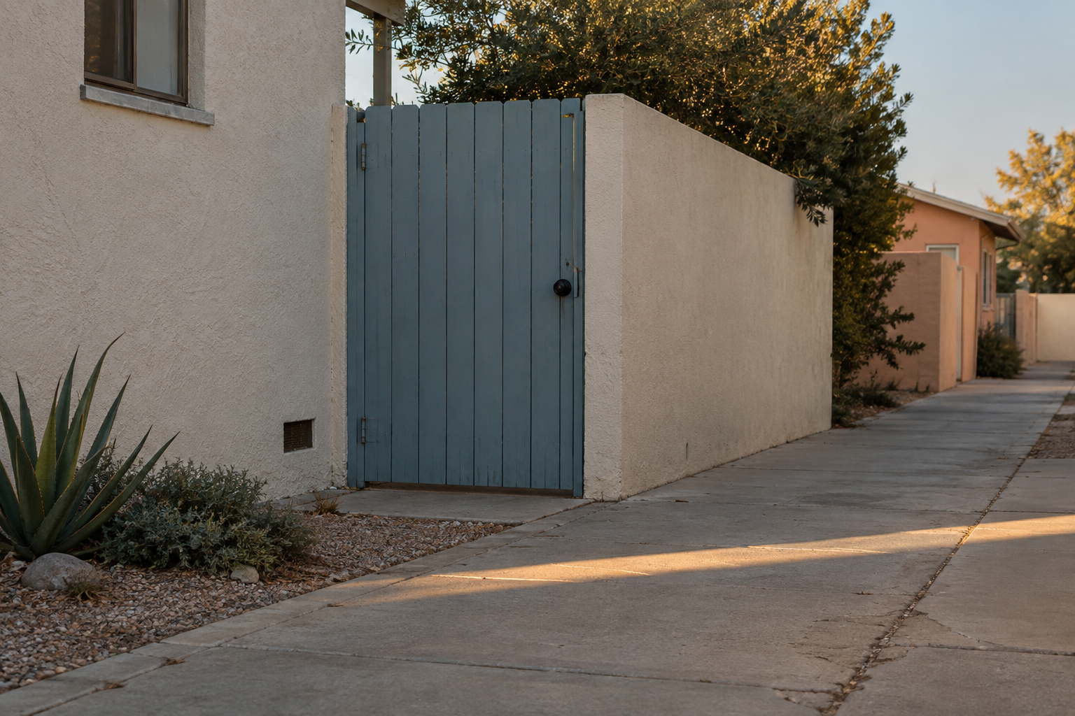 A quiet pedestrian passage with a dusty blue gate and a narrow stripe of late-afternoon light.